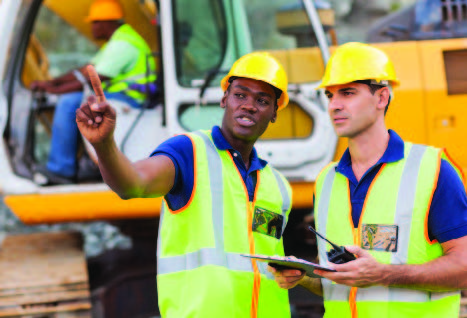 A group of men wearing hard hats, yellow vests and reflectors, man driving heavy equipment in background.