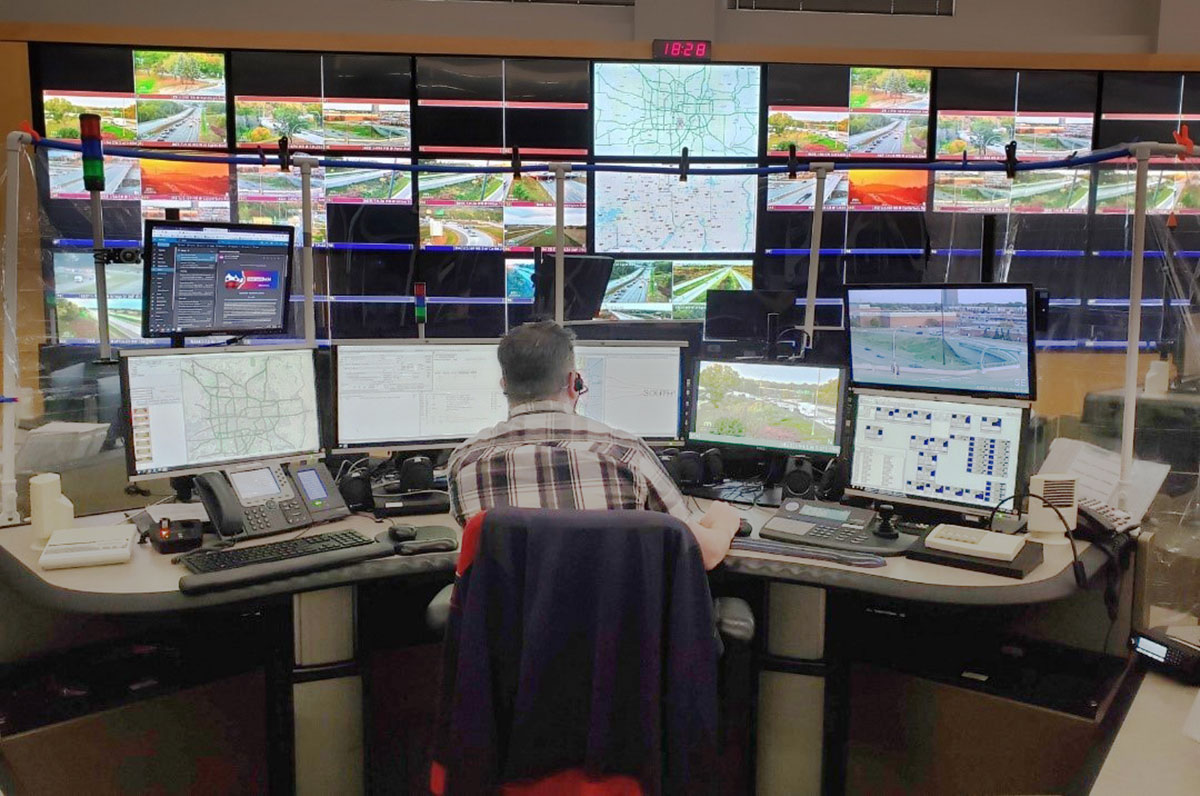 Man, at center, seen from back, sits at large desk surrounded by seven computer monitors. In background, back wall is made up of additional video monitors depicting CCTV feeds and digital maps of roadways