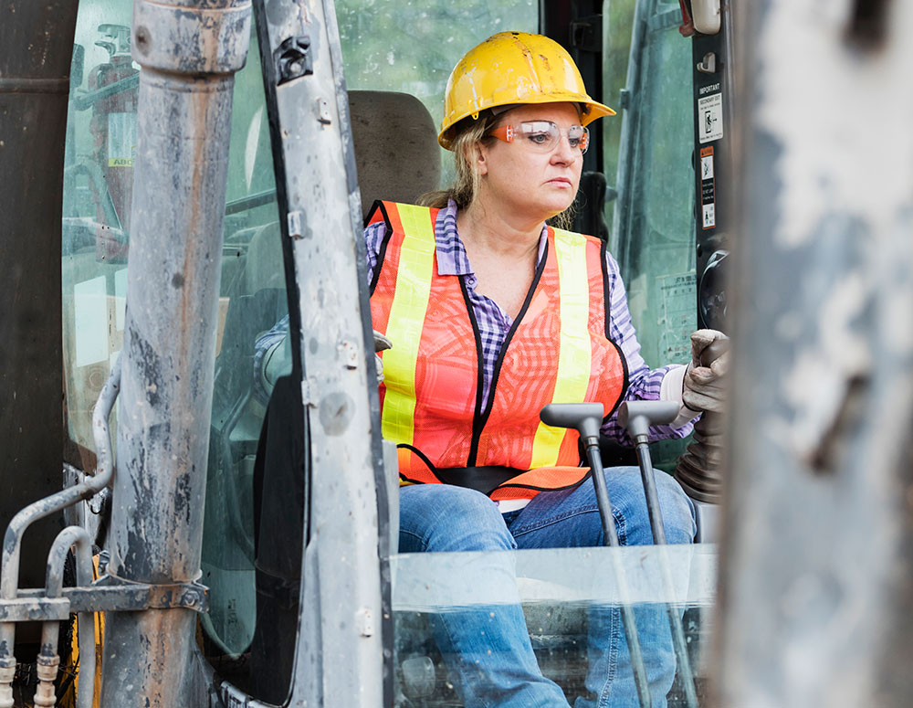 A woman operating heavy machinery wearing hard hat, safety glasses, orange vest and reflectors.