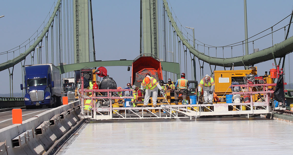 A large piece of equipment extends across a bridge deck with workers on top of a walkway. The equipment is installing a UHPC overlay. On other side of bridge, a truck approaches.