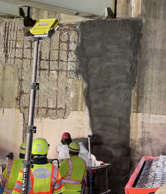 A concrete wall with spalling damage (flaking or breaking-off of surface concrete) and exposed rebar is at rear. A portion of the damage is covered by a dark concrete material, sprayable UHPC. A person is applying the UHPC and others are observing in foreground.