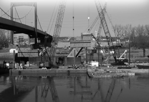 Photograph showing the Missouri River with bridge replacement construction in progress. The contractor on the kcICON project positions forms for the third concrete lift of the downstream river bridge pylon leg. Photo credit to Missouri DOT.