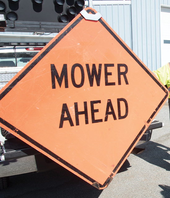 Photo of “MOWER AHEAD” sign attached to back of pickup truck.