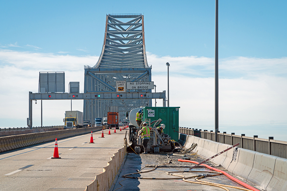 Placement of UHPC on a bridge deck by a work crew of three men in yellow safety vests and hardhats. Cones separate the lane they are working on from a left lane where traffic is moving.