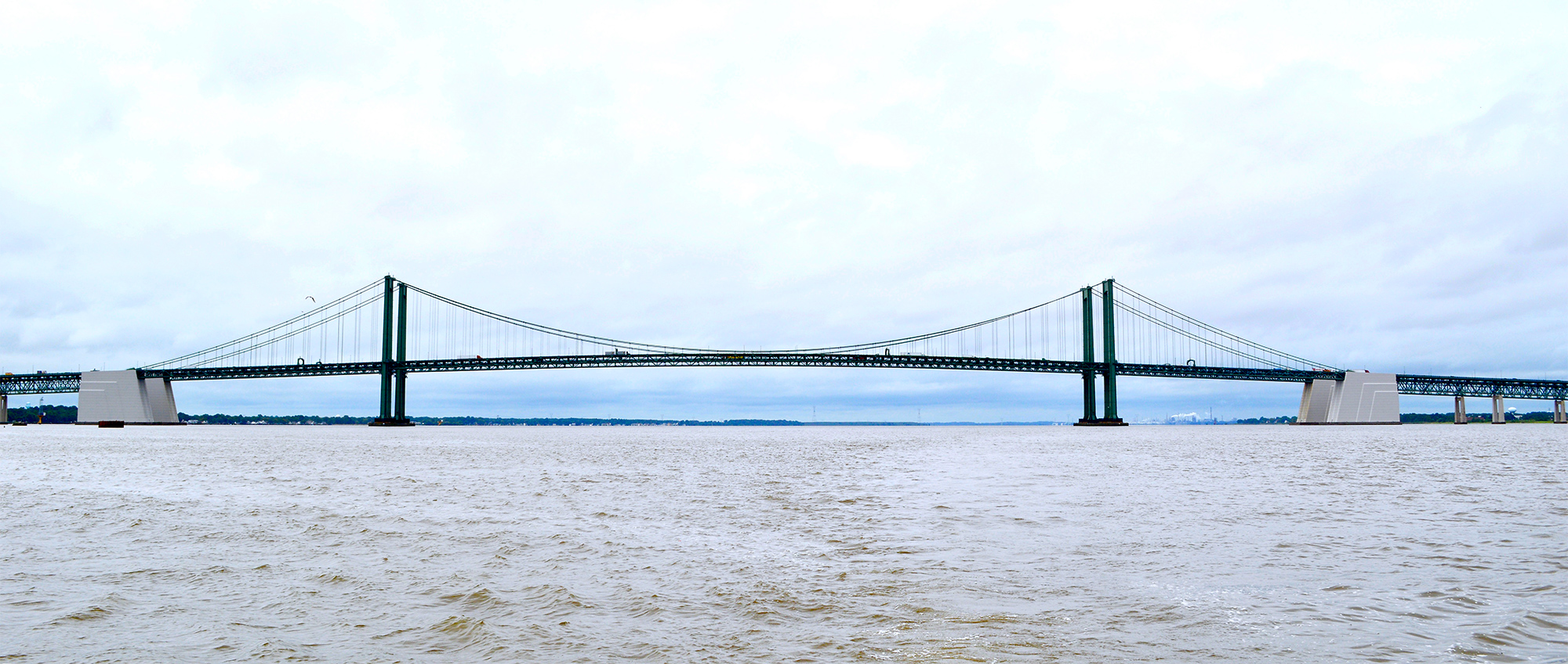 A long-distance view of a twin suspension bridge crossing the Delaware River. The sky is cloudy and the surface of the river is small, choppy waves.