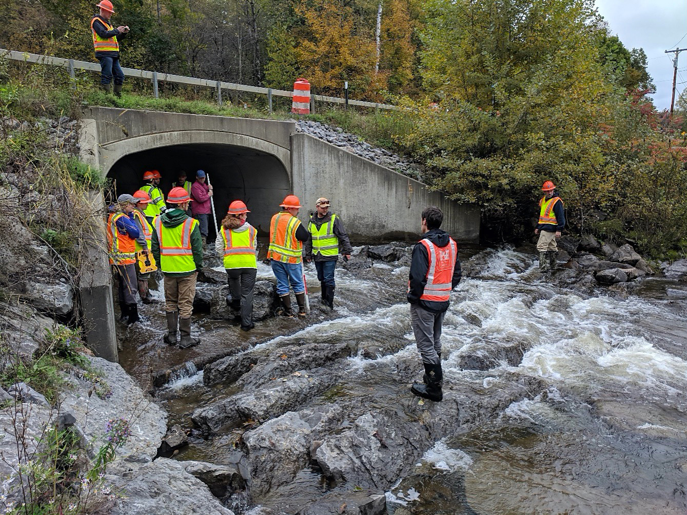 People in wading boots, hard hats, and safety vests stand in a rocky streambed where it exits a culvert.