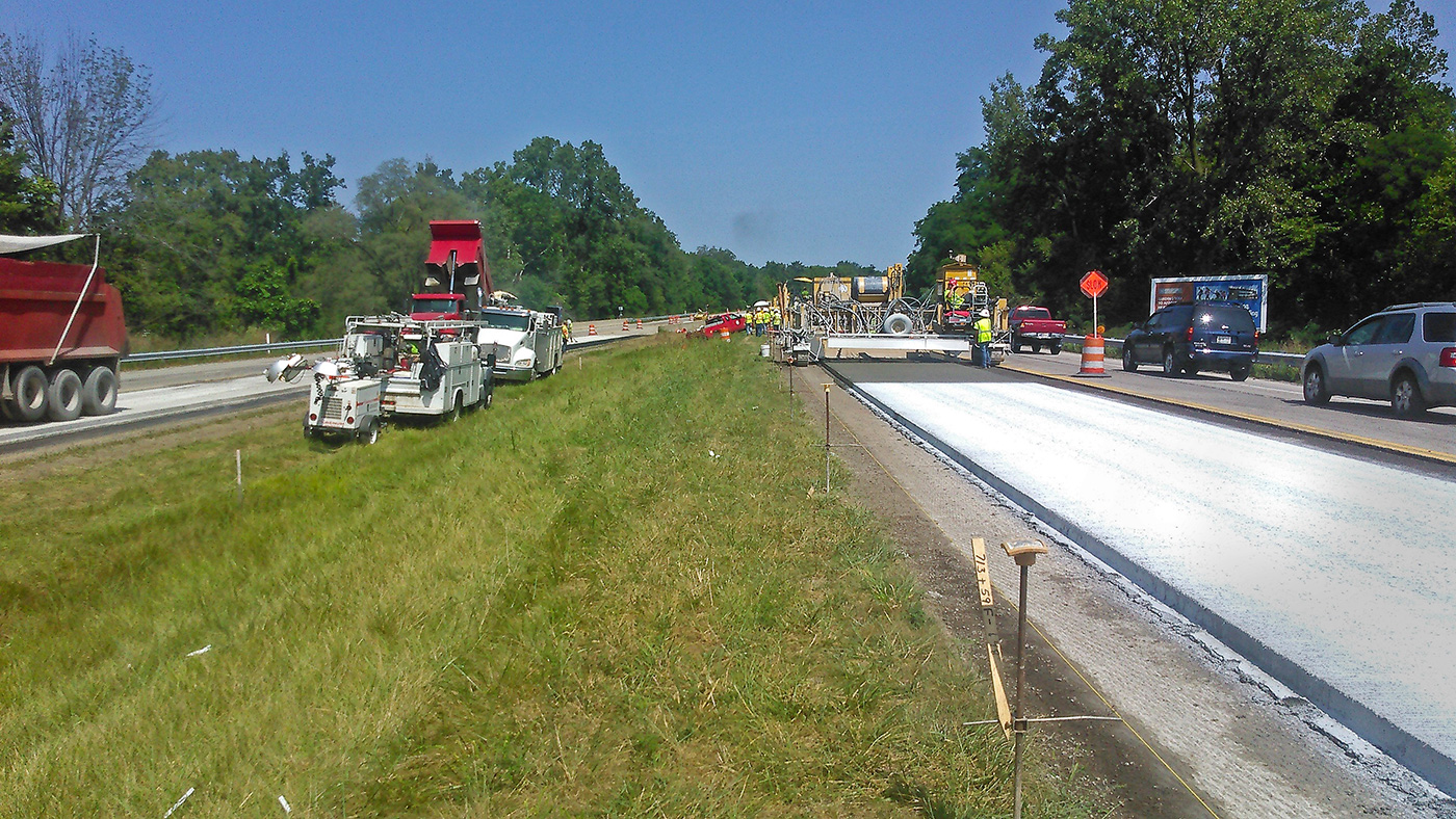 Road construction vehicles are parked in a grass median. One side is closed to traffic and the other has reduced traffic flow as workers operate concrete paving equipment to apply a new overlay on the closed lanes.