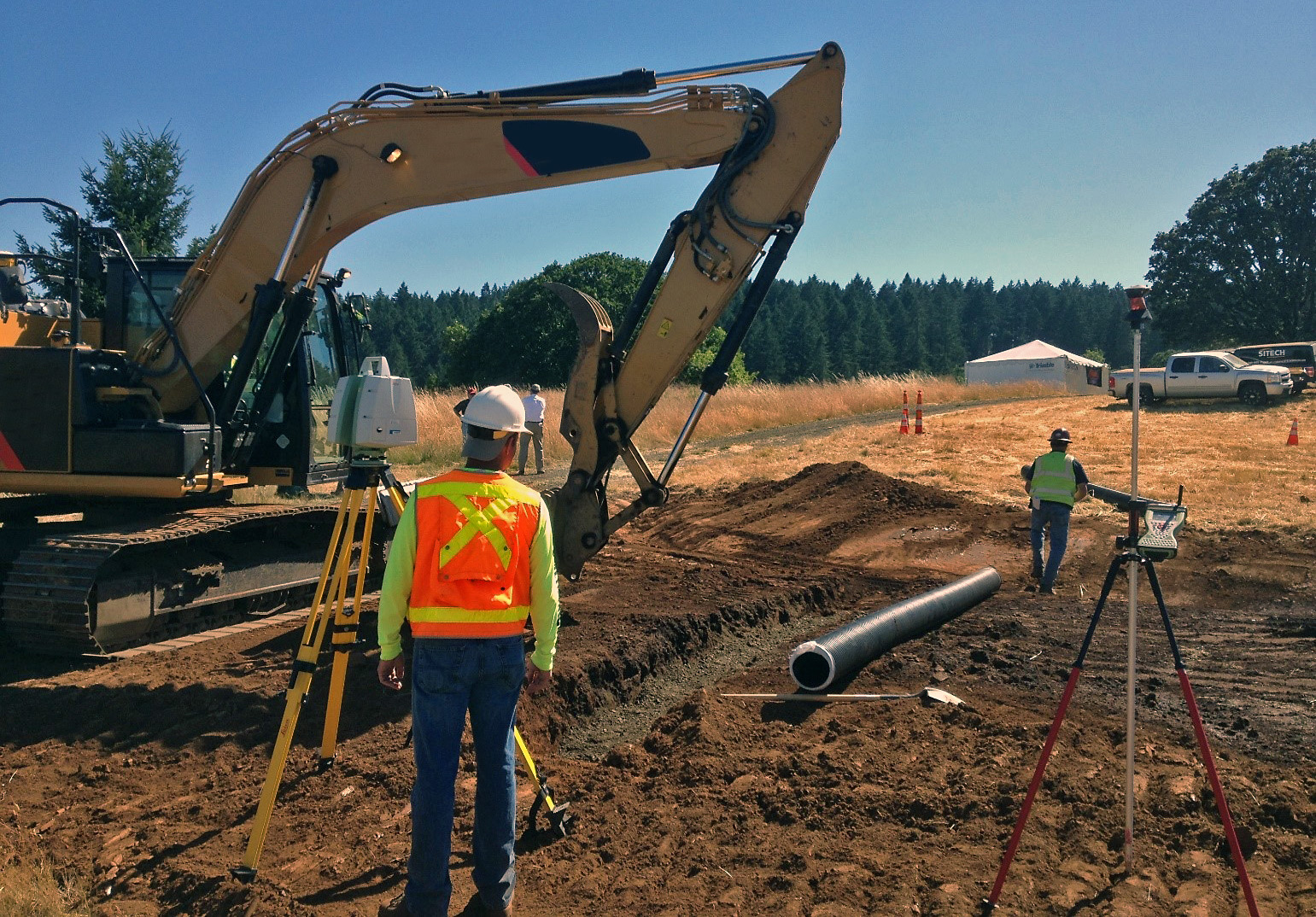 Worker in orange safety vest and hardhat stands near survey equipment and an excavator during pipe installation.