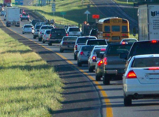 Several cars, a semitruck, a school bus, and a freight truck traveling in close proximity on an interstate highway. Five cars following closely behind each other in the left lane have brake lights activated.