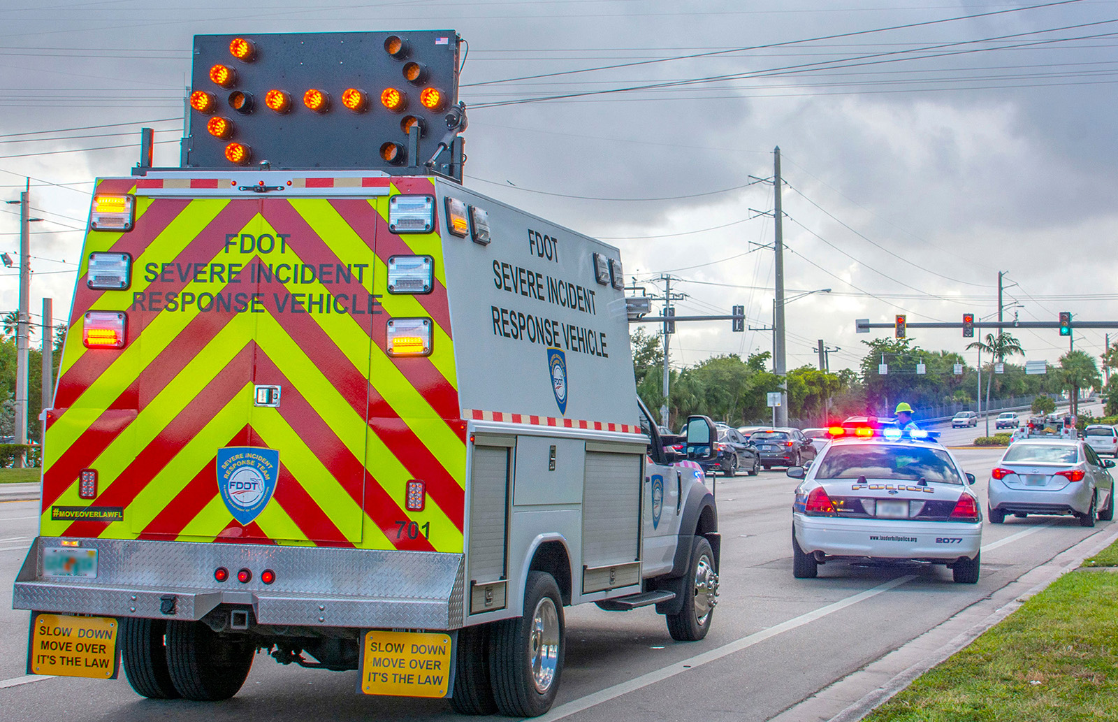 Crash Responder Safety Week. Show your support: #CRSW. November 8–14, 2021. A white truck with yellow and red diagonal stripes and lights on the back and a directional arrow on its roof sits behind a police car with red and blue lights activated. The police car sits diagonally behind a white car stopped on the roadway shoulder.