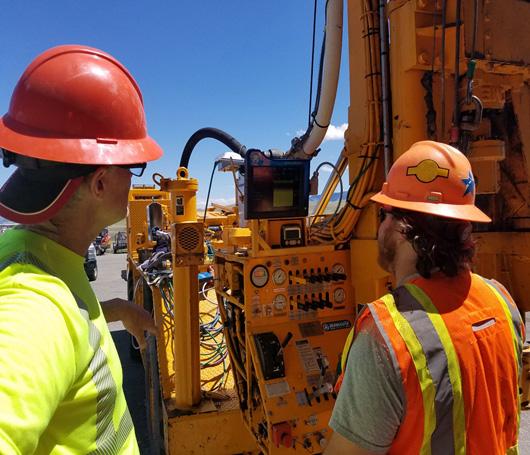 ATwo men wearing safety vests and hardhats look at a digital readout from the instruments installed on a drill rig.