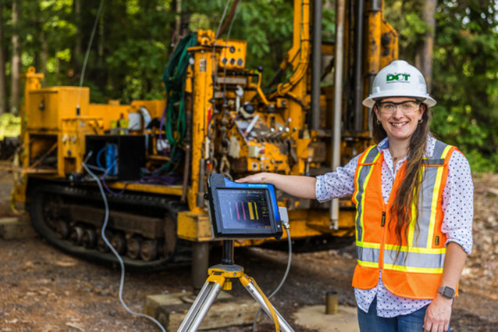 A woman wearing an orange safety vest and white hardhat faces the camera while standing next to an instrument mounted on a tripod with digital readout displayed. A tracked drilling rig sits idle in the background.