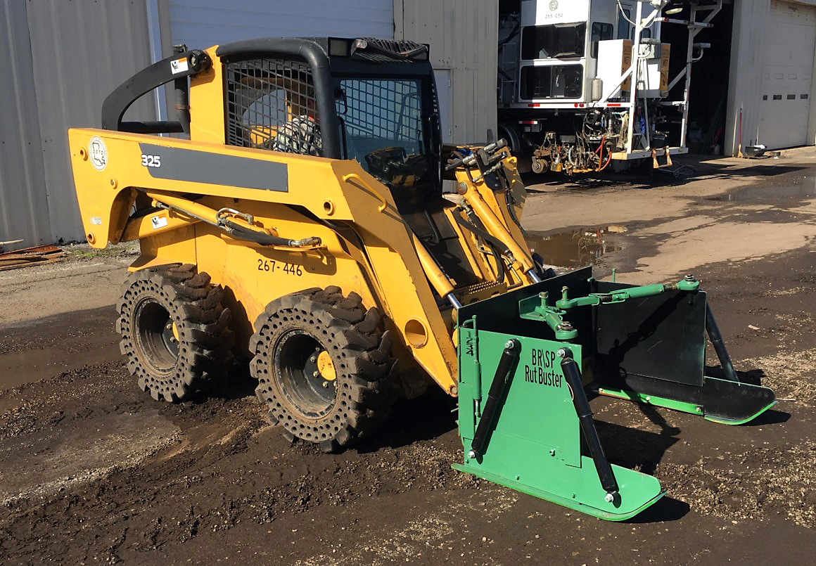 A yellow skid steer loader fitted with a green, u-shaped attachment sits idle on a muddy parking area outside a metal building.