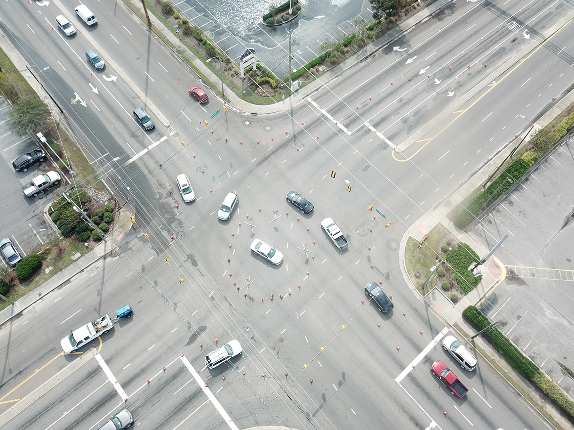 An aerial photo of a four-way intersection with a police car parked in the middle. The police car is surrounded by a circle of orange traffic cones. Additional traffic cones form lanes that direct approaching cars into the traffic circle.