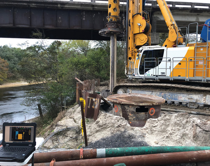 drill rig with monitor taking measurements of the surface next to a girder bridge over water.