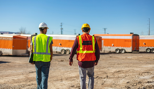 Two workers with florescent vests and hardhats talking.  Picture from behind them.