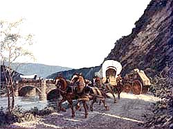 A covered wagan moves down a trail after crossing a stone bridge.