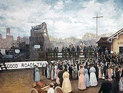 A crowd in Victorian clothing gathers at a train station to meet the 'Good Roads Train.'