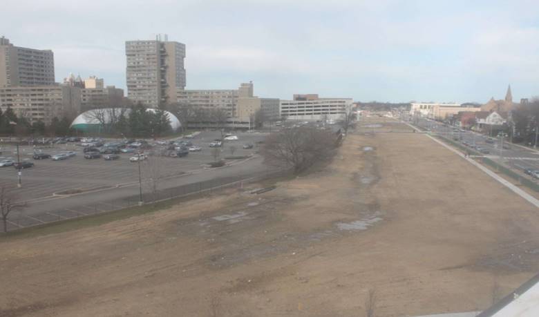 This is an aerial or elevated view of the Inner Loop project area showing roughly six acres of undeveloped land with a roadway on one side and a parking lot and development on the other. This land will eventually be converted into usable space for pedestrians, bicyclists, and others.
