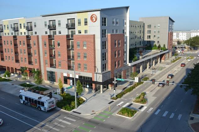 Intersection with traffic signals, parking in front of an apartment building with retail on the ground level, medians for pedestrians to take refuge, a two-way bike lane, and a bus stop. 