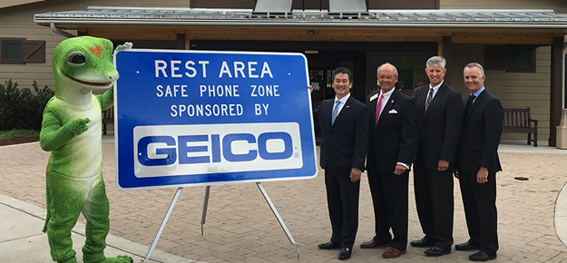 the Geico gecko with  GEICO Regional Vice President John Pham; NCDOT Division 4 Board Member Gus. H. Tulloss; FHWA Division Administrator John Sullivan; and NCDOT Chief Engineer Tim Little next to a rest area sponsorship sign that reads, "Rest Area Safe Phone Zone Sponsored by GEICO".