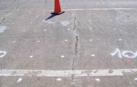 A photograph showing a concrete bridge surface with cracks, an orange traffic cone, and spray-painted markings on the cement is shown. 