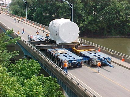 The photograph features a bridge spanning a body of water. Trees appear on each side of the body of water. A very large piece of machinery is supported on two trailers and is crossing the bridge, with the help of numerous construction workers clothed with safety vests and hard hats.