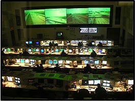 Picture 1. Graphic. The picture shows a Traffic Management Center with several rows of people at desks with monitors and several large wall-mounted screens depicting live roadway conditions.