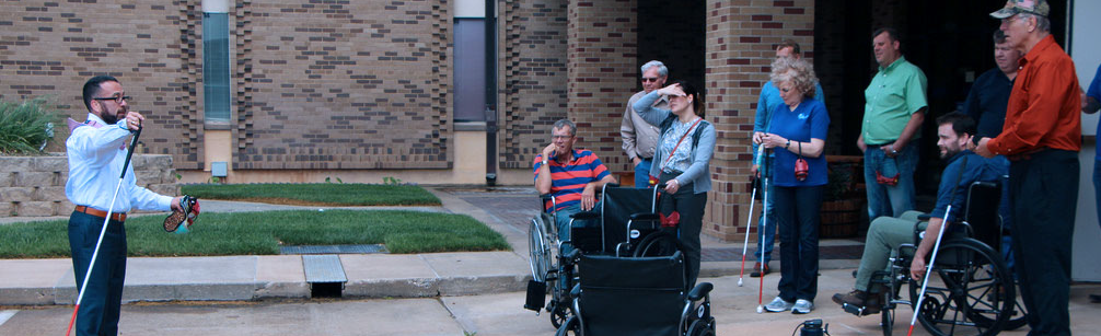 ADA class being conducted outside a building. Instructor Patrick Gomez points while holding a white cane as he addresses a class of 8 individuals using canes and wheel chairs.