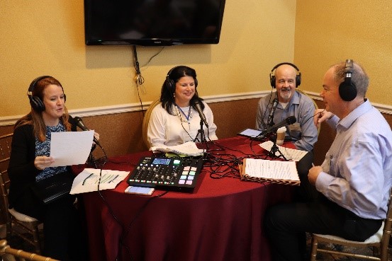 4 people at a table with sound equipment, recording a podcast.