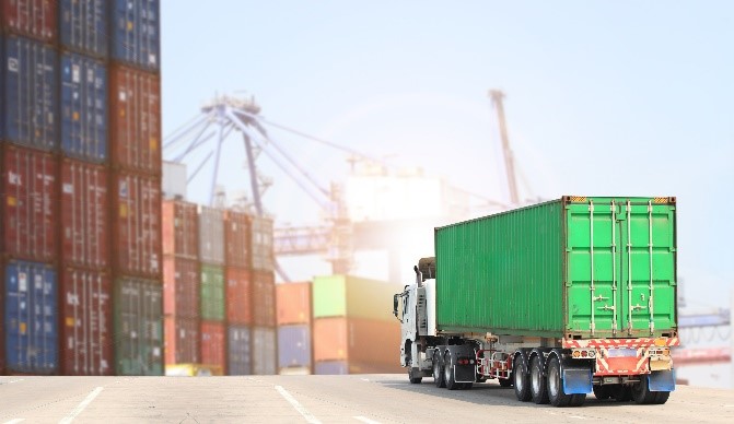 A green truck driving on road with freight containers in background.