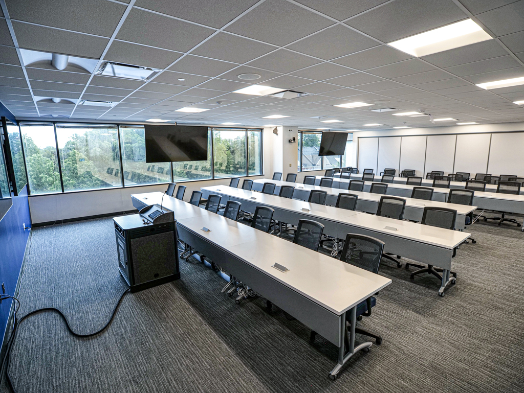 Large training room with 5 rows of tables with 8 chairs per row and screens around the room.