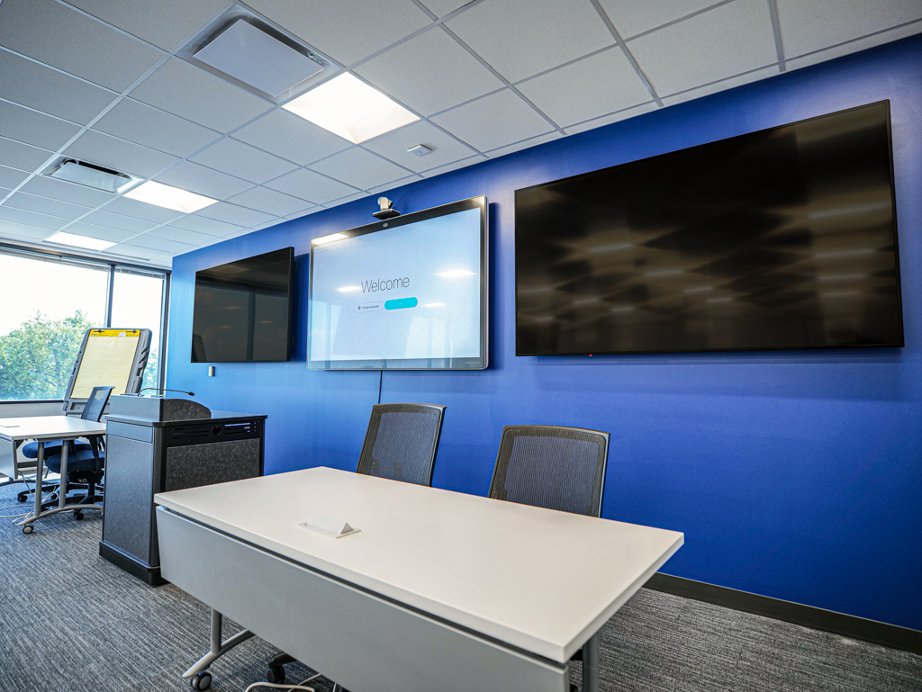 A training room with a blue wall, 3 screens, a podium and a table with chairs in the front of the room.