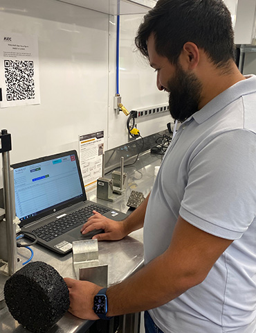 Man inside asphalt trailer laboratory running an asphalt sample preparing to load it into a device to test for rutting potential of the asphalt mix.