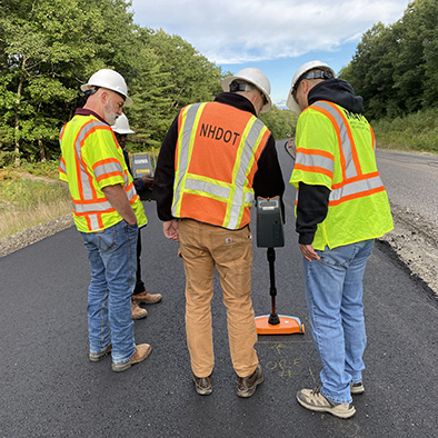 4 men on an active asphalt paving project looking down at the MIT-Scan device which is measuring in-situ pavement thickness.