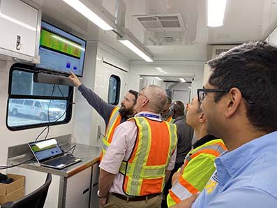 Group of men standing in the interior of a trailer, looking at a computer screen which is showing temperature data on the pavement directly behind a paver.
