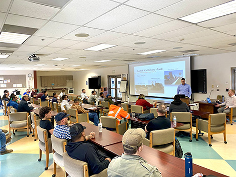  A group of individuals seated in a classroom setting, facing a presenter standing in front of a screen displaying a PowerPoint presentation.