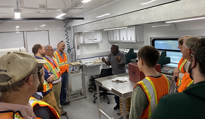 Man inside the interior of a mobile asphalt laboratory who is explaining about the lab to a crowd of visitors.