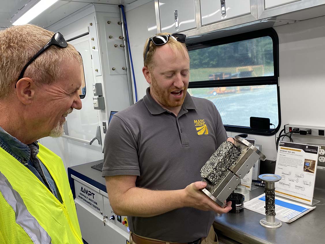 A man showing another man an asphalt specimen that is glued to a metal plated jig that is used in the Overlay Test.