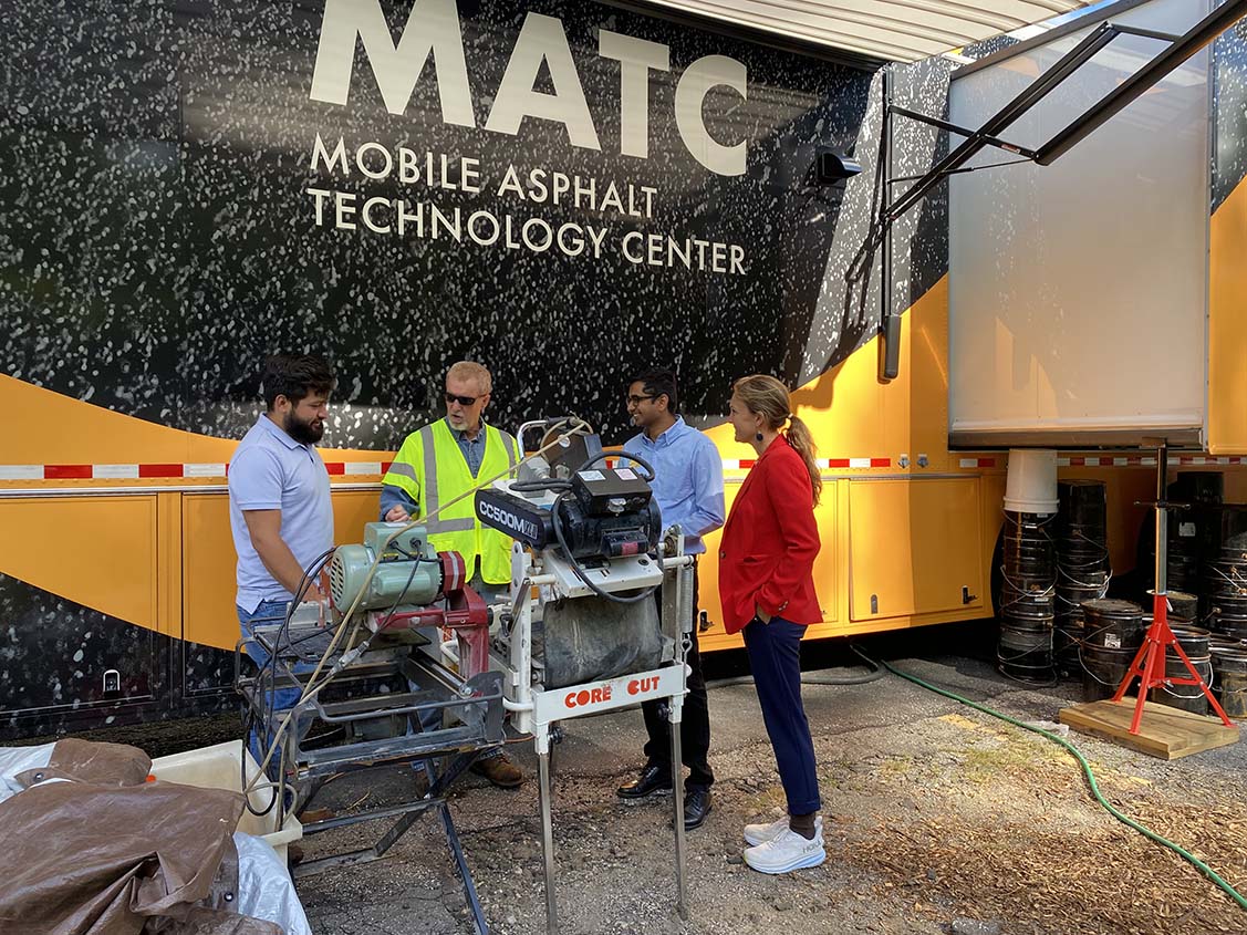 Three men and a woman standing in front of the exterior of the FHWA Mobile Asphalt Technology Center and looking at the saw equipment set up for asphalt core specimen fabrication