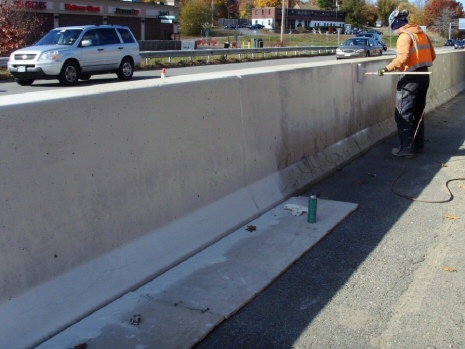 This image shows a worker applying a white-looking paint on a section of concrete barriers using a paint roller. Half of the barriers shown in this image have been painted white.
