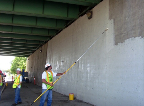 This image shows a worker applying a white paint to a large concrete wall located under a bridge. The worker is using a long paint roller to apply the material.