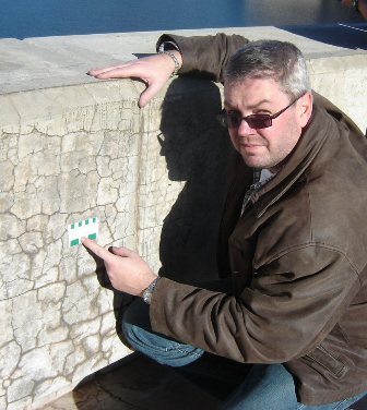 Photograph of editor, Michael Thomas, kneeling next to a concrete barrier exhibiting dark cracks and holding a crack comparator card