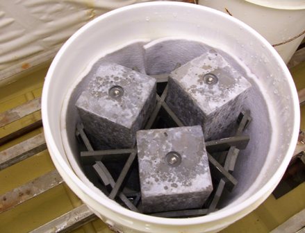 This photo shows a white 5-gallon bucket with three wet rectangular shaped concrete specimens. The specimens are laid out vertically and have embedded steel studs at the ends. The specimens are held in place by a plastic form inside the bucket. There is a white cloth inside the perimeter of the bucket, and there are water droplets inside the bucket.