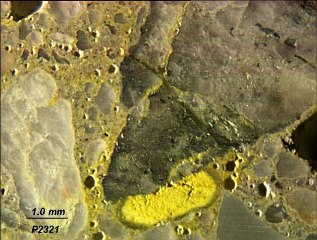 Figure 6. Close-up of Figure 5 showing positive indication for potassium in void-filling deposit along crack in aggregate and in paste around aggregate. This is a close-up picture of the surface of a core extracted from a damaged bridge deck treated with sodium coblatnitrite. Large aggregate particles are visible. Next to one of the large aggregate particles is a yellowish-stained material in a void particle. The rest of the mortar around the aggregates shows a yellow hue. 