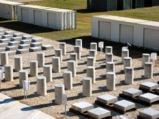 This image shows an aeiral view of several concrete samples standing on a field of gravel. The samples near the top left of the image are rectangular, which are representing pavement slabs. The samples near the middle of the image are cylindrical, which are representing highway columns. The samples near the bottom right of the image are flat and square, which are representing bridge decks.
