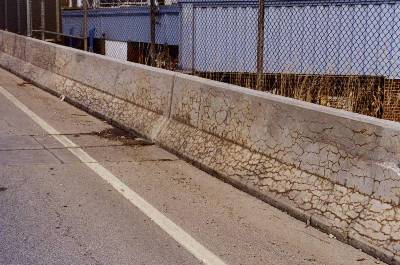 Photograph of a concrete barrier wall along the edge of a road. Severe map cracking on the barrier walls are visible and brown in color. 