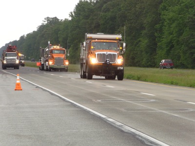 This image shows three large truck vehicles in procession along a section of highway. The first truck is spraying water from its rear, while a second truck follows directly behind.