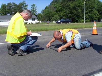 This image shows two men on a section of pavement. One man is crouching down with a clipboard while the second man is holding a length comparator and is measuring the distance between two fixed points on the pavement.