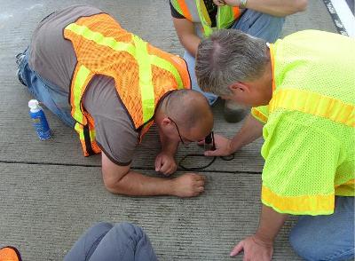 This image shows four people on a section of pavement. One man is crouching down and is looking directly at the surface of the pavement with a hand-held magnifier.